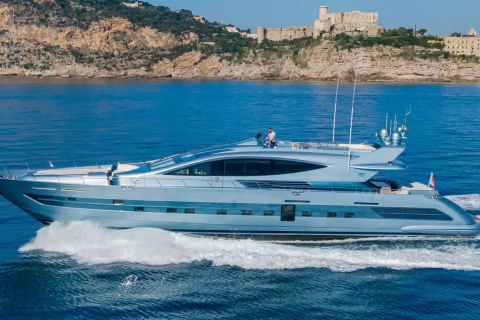 A sleek, modern yacht speeds across the azure waters of a coastal region, with a rocky, cliff-lined shore and buildings visible in the background.