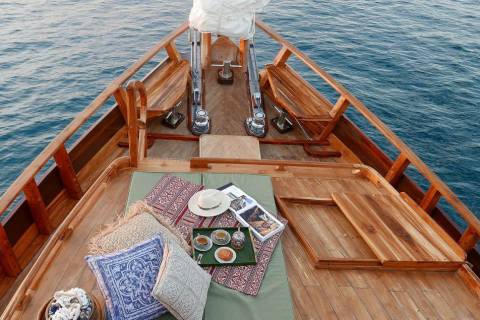 Relax on the wooden deck of a boat with cushions, a hat, and a tea set, surrounded by the calm blue sea.