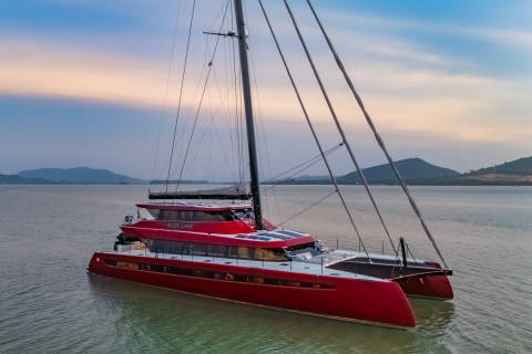 A striking red catamaran named "Crazy Love" glides through calm waters at sunset, its sails furled against a pastel sky.