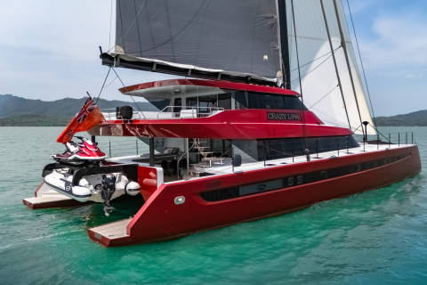 The striking red catamaran "Crazy Love" glides through turquoise waters, its sails catching the breeze under a bright sky.