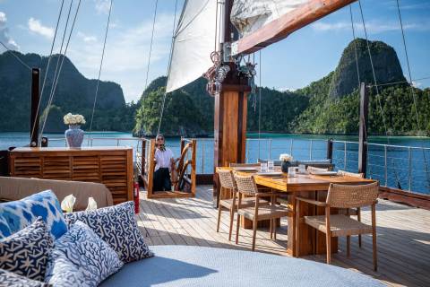 A woman pauses on a wooden deck as a gentle breeze fills the sails of a traditional schooner navigating turquoise waters.