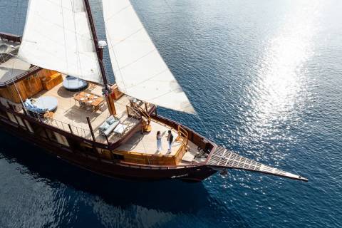 Two people stand on the deck of a luxurious wooden schooner sailing on a sparkling blue sea.