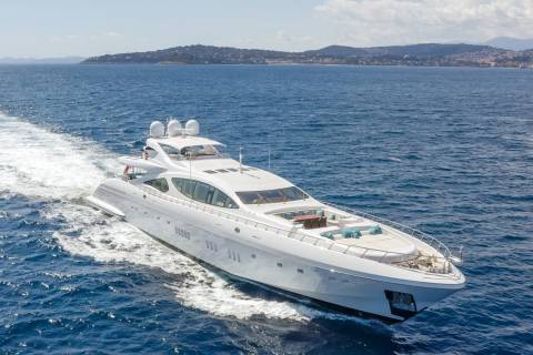 A luxurious white yacht glides through the deep blue sea, leaving a foamy wake behind as it approaches a distant, verdant coastline.
