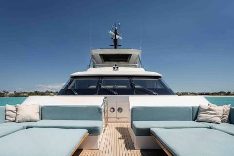 Luxurious light blue seating and plush pillows adorn the deck of a sleek white yacht cruising turquoise waters near a sandy coastline.