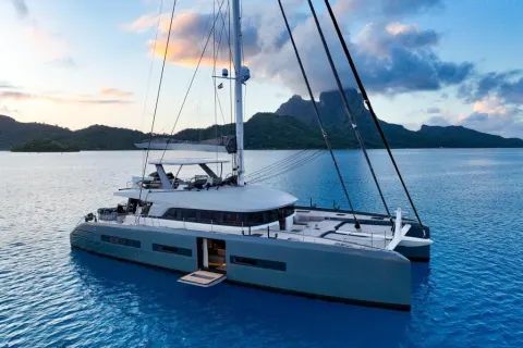 A large, modern catamaran sailboat floating on a calm, blue ocean with lush, mountainous islands in the background under a colorful, cloudy sky.
