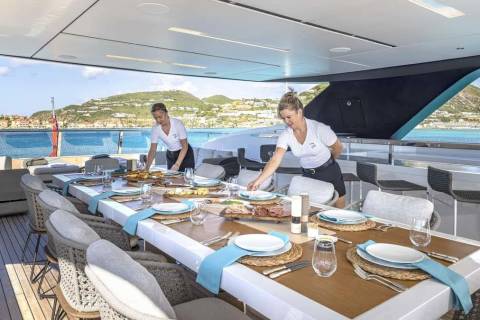 Two servers prepare a lavish outdoor dining table on a yacht overlooking a picturesque coastal town.