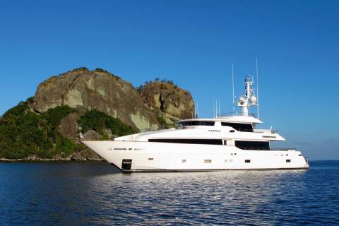 A luxurious white yacht glides through calm blue waters near a lush, rocky island under a clear sky.