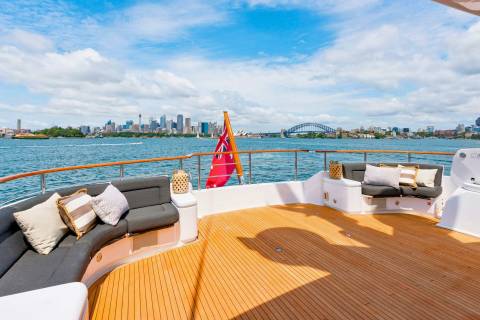 A luxurious yacht deck offers stunning views of the Sydney skyline and Harbor Bridge under a bright blue sky.