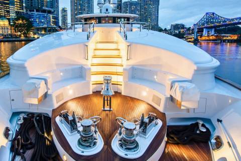The bow of a luxurious yacht is illuminated at dusk, with city lights and a bridge reflecting on the water.