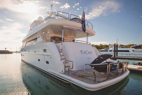 A luxurious yacht named "EnCore" is docked in a serene harbor under a bright, cloudy sky.