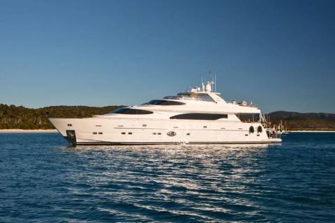 A luxurious white yacht cruises on calm blue waters near a lush, tree-covered coastline under a clear sky.