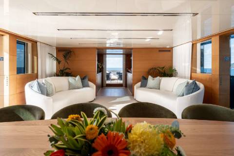 A spacious and modern living room with comfortable white sofas, a wooden dining table, and vibrant floral arrangements, surrounded by large windows offering a scenic view.
