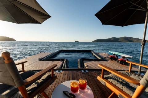 A wooden deck with lounge chairs and umbrellas overlooks a serene body of water with distant islands in the background.