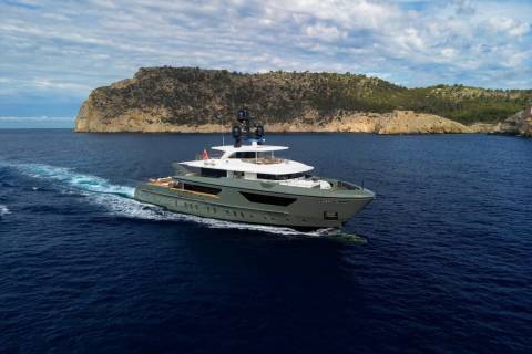 A sleek, modern yacht glides through the deep blue waters near a rugged, tree-covered island under a cloudy sky.