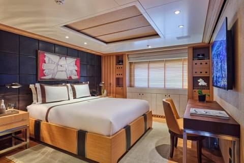 A luxurious yacht cabin features a king-sized bed, modern artwork, and a sleek desk with a view of the ocean on the television.