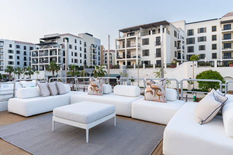 Luxurious white outdoor seating with flamingo-patterned pillows offers a stylish retreat against a backdrop of modern waterfront buildings.