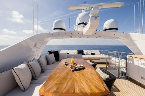 A luxurious yacht deck offers a sun-drenched dining area with plush seating and a vibrant fruit platter overlooking the serene blue ocean.