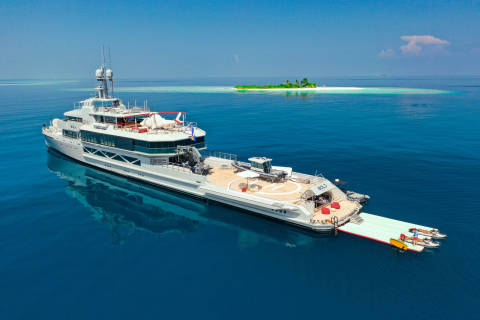 A luxurious yacht named Bold anchors in the clear blue waters near a small tropical island.