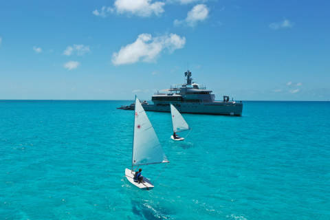 Two small sailboats glide across turquoise waters near a large yacht under a bright blue sky.