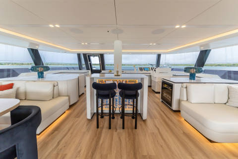 A modern and spacious interior of a yacht or boat, featuring a sleek and minimalist design with a kitchen area, bar stools, and comfortable seating arrangements, all surrounded by large windows offering a scenic view of the water in the background.