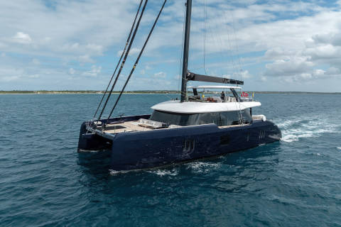 A large black catamaran sailboat is sailing across a body of water surrounded by a scenic coastal landscape under a partly cloudy sky.