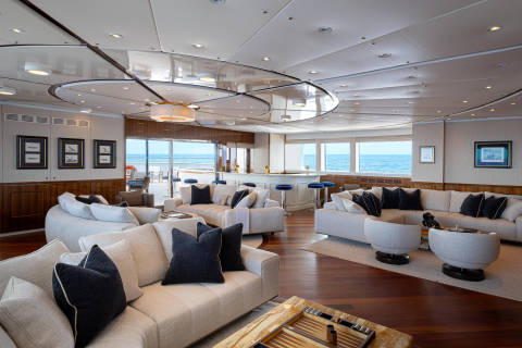 A spacious and luxurious lounge area with plush white and black sofas, a curved ceiling with recessed lighting, and large windows offering a scenic view of the water in the background.