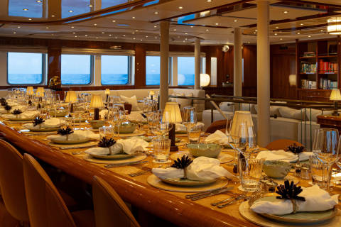 A luxurious dining room with a panoramic view of the ocean, featuring an elegantly set table with fine china, glassware, and decorative elements.