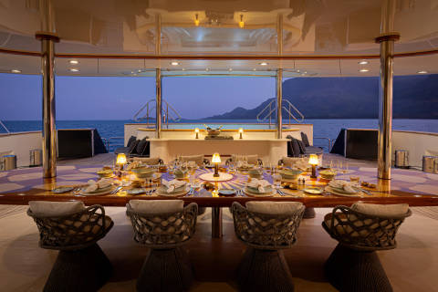 A luxurious dining area on a yacht, with a table set for a formal meal and surrounded by large windows offering a breathtaking view of the ocean and mountains in the background.