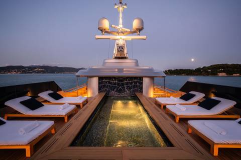 A luxurious yacht with a sleek design, featuring a central structure with a cross on top, surrounded by lounge chairs and a pool-like area, set against a scenic backdrop of a body of water and a night sky.