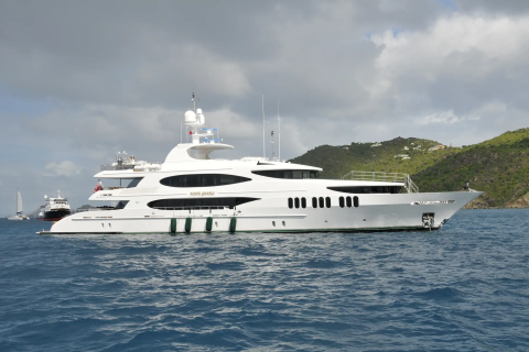 A large white yacht with green trim sails on blue water in the foreground, while a lush green island with scattered buildings and a cloudy sky form the background.