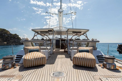 Two striped lounge chairs sit on the wooden deck of a yacht, with a calm blue sea and a tree-covered island in the background.