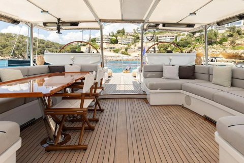 A spacious yacht deck with a dining table and lounge seating in the foreground overlooks a picturesque coastline with buildings nestled on a hill in the background.