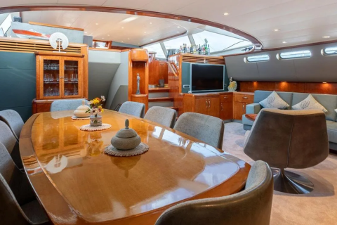 A polished wooden dining table with chairs in the foreground is set against a backdrop of a luxurious yacht interior featuring a television, sofas, and wooden cabinetry.