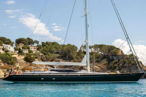 A sleek, dark green yacht floats on turquoise water in the foreground, with a lush, tree-covered hillside dotted with white buildings in the background.