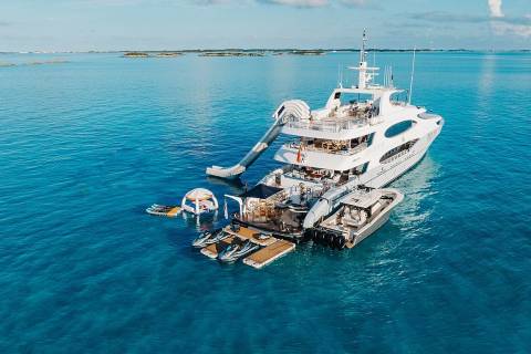 A large white yacht with a slide and various water toys is anchored in clear turquoise water with a distant island on the horizon.
