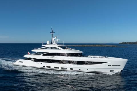 A sleek white yacht cruises through calm blue waters with a distant, tree-lined coastline under a clear sky.