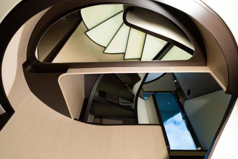 A modern spiral staircase with cream-colored steps and dark brown railings curves upwards towards a bright sky visible through a window in the background.
