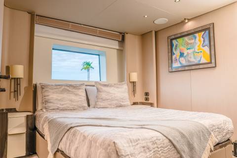 A neatly made bed with plush pillows and a light gray throw occupies the foreground, while a framed abstract painting hangs on the wall above a nightstand in the background, with a window revealing a palm tree and sky.
