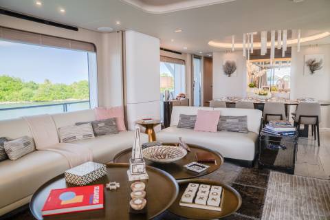 A modern yacht living room with plush white sofas and coffee tables in the foreground, and a dining area with a long table and chairs in the background.