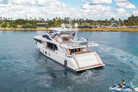 A luxurious white yacht cruises on calm blue water in the foreground, with a jet ski trailing behind, while palm trees and a distant cityscape line the background.