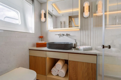 A modern bathroom features a dark basin on a wooden vanity in the foreground, with a large mirror and wall sconces reflecting the bright, clean background.