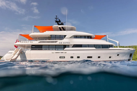 A large white yacht with orange awnings floats on calm blue water in the foreground, with a lush green island and a bright blue sky in the background.
