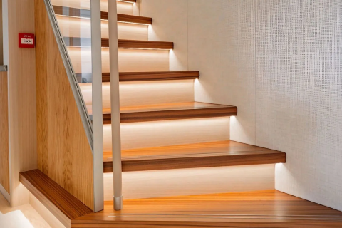 A modern wooden staircase with illuminated steps ascends against a textured white wall, with a fire alarm visible on the left.