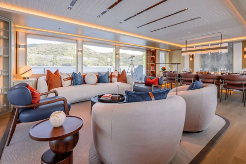 Plush armchairs and a sectional sofa with colorful pillows fill the foreground of a luxurious yacht lounge, while a large dining table and panoramic windows revealing a scenic coastal landscape form the background.