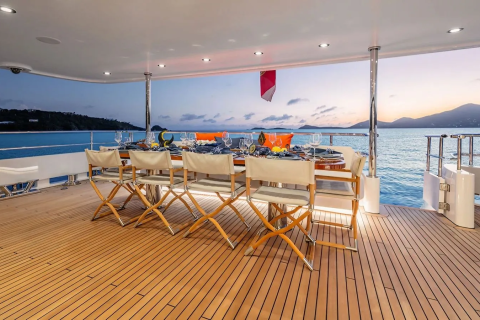 A beautifully set dining table with chairs on a yacht's deck overlooks a serene ocean and distant islands at dusk.