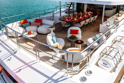 A luxurious yacht deck with comfortable seating and a dining table in the foreground, set against a backdrop of calm blue water.