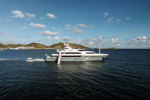 A large white yacht with a slide is anchored in the deep blue sea, with a hilly coastline dotted with white buildings in the background under a cloudy sky.