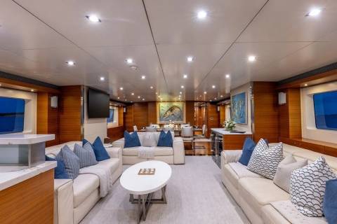 A spacious yacht interior with plush white sofas and blue accent pillows in the foreground, and a dining area with artwork and large windows overlooking the sea in the background.