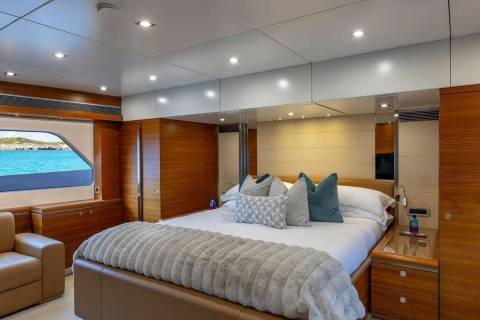 A luxurious yacht bedroom with a large bed and plush pillows in the foreground, and a window overlooking a turquoise ocean and distant islands in the background.