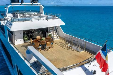 A luxurious yacht with an outdoor dining area in the foreground sails on a calm, turquoise ocean under a clear sky.